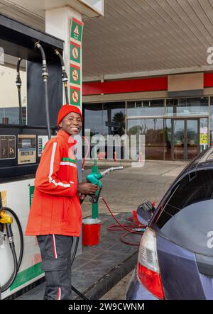 Tankstellenwart, afroamerikaner in roter Uniform mit der Düse in der Hand, die ein Auto betankt Stockfoto