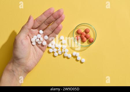 In der Handfläche liegen einige Pillen, während andere verstreut sind und mit einer Petrischale und mehreren orangen Pillen auf gelbem Hintergrund verziert sind. Stockfoto
