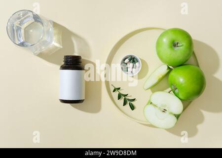 Eine runde Keramikschale mit grünem Apfel und Pillen auf einer Kappe, eine Arzneiflasche und eine Glasschale auf beigefarbenem Hintergrund. Grüne Äpfel haben m Stockfoto