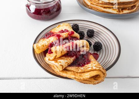 Teller mit köstlichen frisch gebackenen Crepes mit Marmelade und Beeren auf einem weißen Holztisch. Traditionelles Essen für den Urlaub in Russland, um den Winter zu genießen Stockfoto