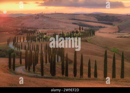 Toskanische Sonne untergeht auf einer Zypressen-Baumstraße und rustikales Bauernhaus in der malerischen sanften Landschaft der Toskana, Italien. Stockfoto
