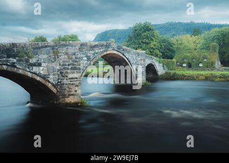 Alte römische Brücke Pont Fawr über den River Conwy in Richtung eines mit Efeu bedeckten Steinhauses aus dem 15. Jahrhundert im walisischen Dorf Llawnrst, Nordwales Stockfoto