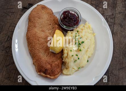 Schweineschnitzel nach Wiener Art aus Strohschwein - in knuspriger Panierung serviert mit hausgemachtem Kartoffelsalat, Zitrone und Preiselbeeren auf Keramikplatte. Draufsicht, Stockfoto