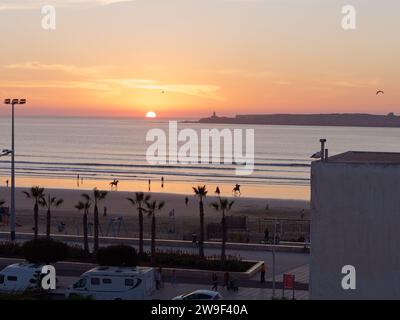 Erhöhter Blick bei Sonnenuntergang von Pferdereitern an einem Sandstrand, während der Sonnenuntergang sich im Wasser spiegelt. Essaouira, Marokko. Dezember 2023 Stockfoto