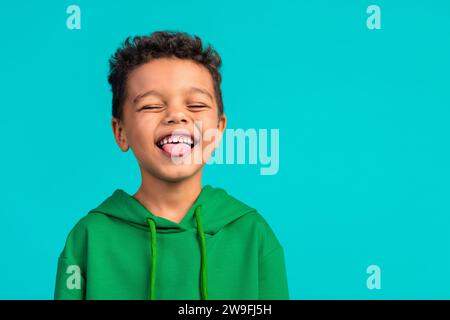 Porträt des überglücklich lvoely kleinen Jungen mit lockigen Haaren tragen grünen Hoodie zeigen Zunge haben Spaß isoliert auf türkisfarbenem Hintergrund Stockfoto