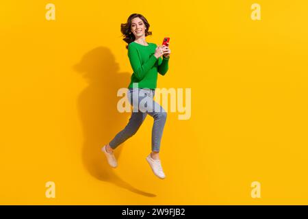 Ganzkörper-Foto von fröhlicher junger energetischer Dame grüne Pullover-Jeans, die mit Handy auf gelbem Hintergrund isoliert sind Stockfoto