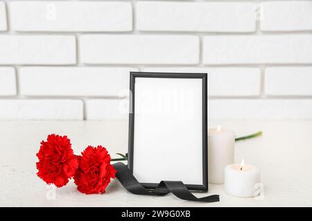 Leerer Trauerrahmen, Nelkenblumen und brennende Blumen auf dem Tisch in der Nähe einer hellen Ziegelwand Stockfoto