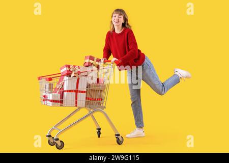 Junge Frau mit Einkaufswagen voller Weihnachtsgeschenkboxen auf gelbem Hintergrund Stockfoto