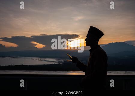 Silhouette eines asiatischen muslimischen Mannes, der ein Smartphone bei Sonnenuntergang hält. Ein Mann mit einem Smartphone unter dem bewölkten Sonnenuntergang. Stockfoto