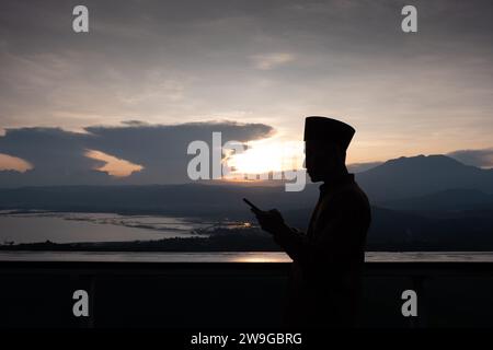 Silhouette eines asiatischen muslimischen Mannes, der ein Smartphone bei Sonnenuntergang hält. Ein Mann mit einem Smartphone unter dem bewölkten Sonnenuntergang. Stockfoto