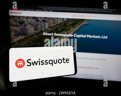 Person mit Handy mit Logo des Schweizer Finanzdienstleisters Swissquote vor der Business-Webseite. Schwerpunkt auf dem Display des Telefons. Stockfoto