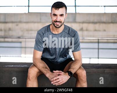 Junger Athlet, der lächelnd sitzt und in die Kamera schaut. Fröhlicher aktiver Läufer, der vom Training im Fitnessstudio zufrieden ist. Stockfoto