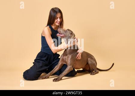 Der Blick von Liebe und Zärtlichkeit. Junge brünette Frau sitzt auf dem Boden und umarmt sich mit ihrem Lieblingstier vor beigefarbenem Studio-Hintergrund. Stockfoto