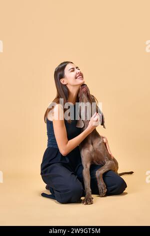 Umarmungen und Küsse, dogfreundlich. Lifestyle. Junge brünette Frau sitzt auf dem Boden und umarmt sich mit ihrem Lieblingstier vor beigefarbenem Hintergrund. Stockfoto