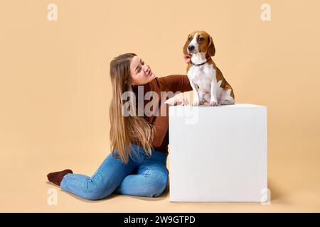 Porträt einer jungen hübschen Frau mit ihrem reinrassigen Beagle, Hund vor beigefarbenem Atelierhintergrund. Vorstellung von Freundschaft, Liebe. Stockfoto