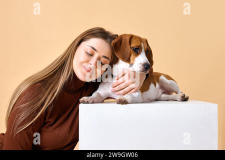 Studio-Porträt von Hund und Besitzer. Junge Frau lächelt und umarmt ihren Lieblingshündchen Beagle vor beigefarbenem Hintergrund. Stockfoto