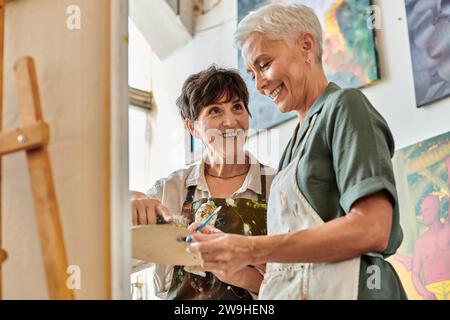 Fröhliche Künstlerinnen mittleren Alters, die während des Meisterkurses im Kunstworkshop Farben auf Palette mischen Stockfoto