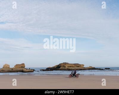 Der Mensch fischt auf einem Felsvorsprung am Rande eines Sandstrandes, während jemand auf einem Motorrad in der Stadt Essaouira, Marokko, sitzt. Dezember 2023 Stockfoto