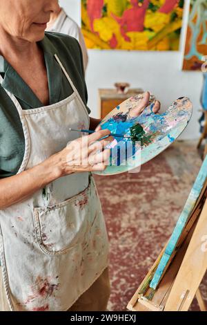 Zugeschnittene Ansicht einer reiferen Künstlerin, die Farben auf der Palette mit Pinsel in der Kunstwerkstatt vermischt Stockfoto
