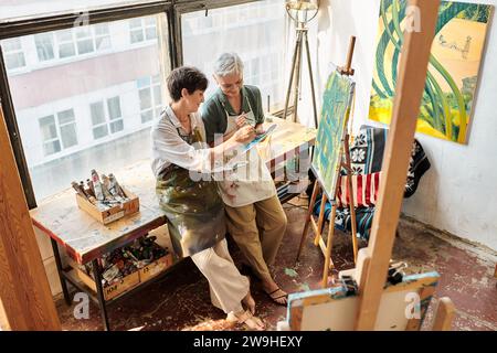 Glückliche Frauen mittleren Alters mischen Farben auf Paletten in der Nähe von Staffeleien im Kunststudio, kreatives Hobby Stockfoto