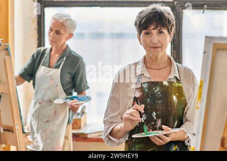 Kreative reife Frau, die die Farbpalette hält und während des Meisterkurses im Kunststudio in die Kamera schaut Stockfoto