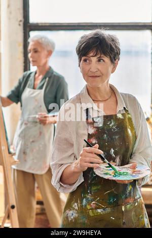 Lächelnde reife Frau, die Farbpalette hält und während des Meisterkurses in der Kunstwerkstatt wegschaut Stockfoto