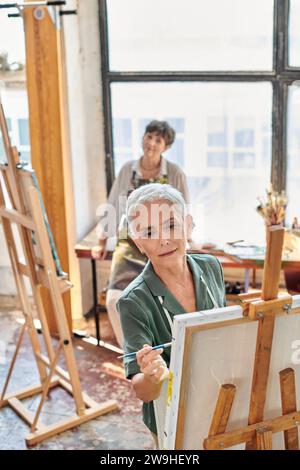 Inspirierte reife Frau in Schürzenmalerei auf Staffelei in Meisterklasse in Kunstwerkstatt, kreatives Hobby Stockfoto