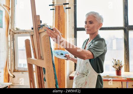 Konzentrierte grauhaarige reife Frau in Schürze, die Staffelei in der Handwerkstatt betrachtet, kreatives Hobby Stockfoto
