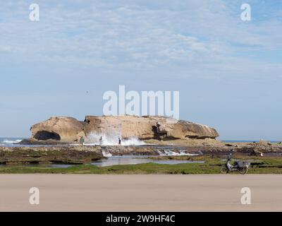 Einheimischer Fischer neben großen Felsvorsprüngen neben einem Sandstrand, während Wellen in Essaouira, Marokko, über sie krachen. Dezember 2023 Stockfoto