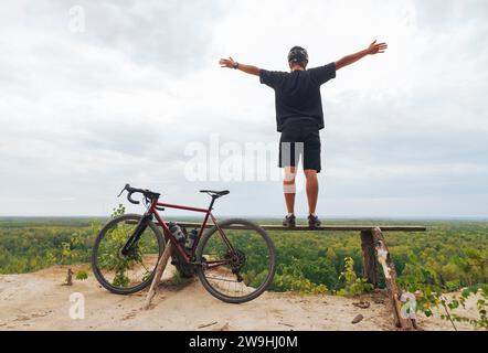 Junger glücklicher Mann mit Fahrrad steht auf einer Klippe und genießt die Aussicht mit ausgestreckten Armen. Stockfoto