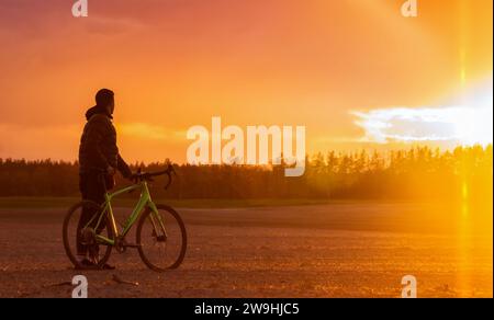 Ein Mann mit einem Schotterrad steht auf einem Feld auf einem wunderschönen Sonnenuntergang Hintergrund. Wunderschöne Landschaft mit Mountainbike. Stockfoto