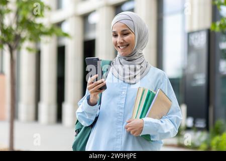 Junge muslimische Frau im Hijab, die außerhalb des Universitätsgeländes läuft, die Studentin lächelt zufrieden mit der App auf dem Telefon, dem Rucksack auf dem Rücken und Büchern in den Händen. Stockfoto