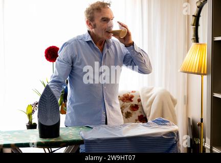 Älterer Rentner macht eine Kaffeepause, während er zu Hause Kleidung bügelt Stockfoto