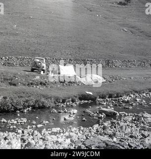 1960er Jahre, historisch, sommerlich und Camping an einem ländlichen Fluss auf einem flachen Stück Land an einer Steinmauer im hügeligen Peak District, England, Großbritannien. Auto der Ära mit Nummernschild UWR 475. Stockfoto