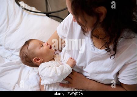 Authentisches Porträt einer lockigen, brünetten jungen erwachsenen Frau, einer glücklichen, liebevollen Mutter, die lächelt und positive Emotionen ausdrückt, während sie stillt und kuschelt Stockfoto