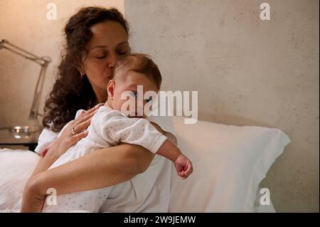 Porträt einer authentischen, schönen lateinamerikanischen Frau im weißen Pyjama, die ihren kleinen Jungen in der Hand hält und sanft kuschelt, zusammen auf dem Bett sitzt Stockfoto