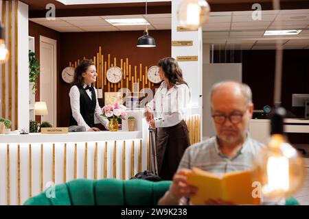 Ältere weibliche Gäste mit Gepäck, die mit der jungen Rezeptionistin an der Rezeption des Luxusresorts sprechen. Ein alter Mann mit einem Roman entspannt sich in der Hotellobby, während eine ältere Frau am Schalter Buchungen macht. Stockfoto
