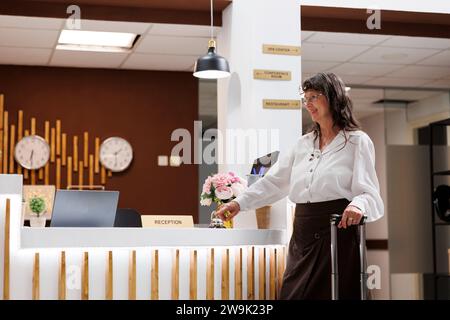 Eine Seniorin erreicht den Meldeschalter und ruft die Metallglocke an, um von den Rezeptionisten des Hotels nach Service zu fragen. Kaukasische ältere Frau, die auf Unterstützung durch das Resort-Personal wartet. Stockfoto