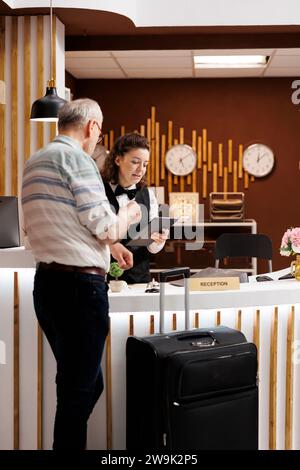 Älterer männlicher Reisender meldet sich am Schalter der Hotellobby an, um einen angenehmen Urlaub zu beginnen. Ein pensionierter Seniorenmann füllt Reservierungsunterlagen mit Hilfe einer netten weiblichen Rezeptionistin aus. Stockfoto