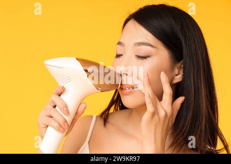 Hübsche asiatische Frau, die einen Fotoepilierer auf ihrem Gesicht vor gelbem Hintergrund benutzt Stockfoto