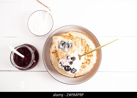 Blick von oben auf eine große Keramikplatte mit Crepes mit Sauerrahm und frischen Beeren. Ein Glas Himbeermarmelade, eine Schüssel Sauerrahm. Stillleben Stockfoto