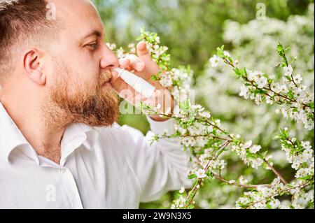 Mann allergisch mit medizinischen Nasentropfen, leidet an saisonaler Allergie im Frühling im blühenden Garten. Schöner Mann, der die laufende Nase vor blühenden Bäumen im Freien behandelt. Frühjahrsallergiekonzept. Stockfoto
