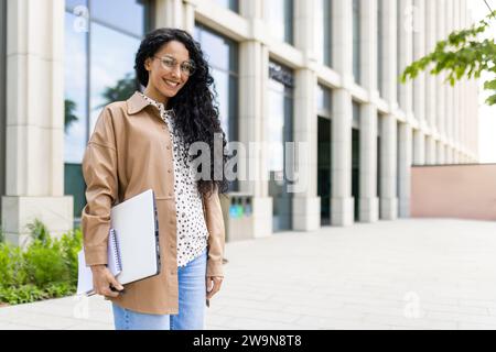 Porträt einer jungen schönen erfolgreichen Geschäftsfrau vor dem Bürogebäude, einer erfolgreichen lateinamerikanischen Frau, die lächelt und in die Kamera blickt, mit einem Laptop in den Händen und Dokumenten steht. Stockfoto