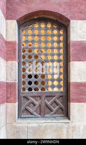 Rustikaler Charme der Mamluk-Architektur, verwittertes Holzfenster mit einem einzigartigen sternförmigen Muster. Das Fenster ist vor einem Hintergrund von alten roten und weißen Ziegelmauern, die einen Hauch von Geschichte hinzufügen Stockfoto
