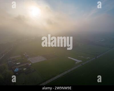 Das Bild ist eine eindrucksvolle Luftaufnahme, die die ätherische Schönheit des Morgengrunds festnimmt, während die Sonne durch den Morgennebel bricht. Ein ländliches Gehöft liegt ruhig, flankiert von ordentlich geteilten landwirtschaftlichen Feldern. Die aufgehende Sonne erzeugt ein warmes Leuchten, das einen beeindruckenden Hinterleuchteffekt und lange Schatten erzeugt. Der Nebel weicht die Details der Landschaft auf und verleiht der Szene eine traumhafte Qualität. Diese ruhige Umgebung ist eine eindrucksvolle Darstellung der Ruhe und Einfachheit des ländlichen Lebens. Ätherische Morgenröte Über Dem Landhaus. Hochwertige Fotos Stockfoto