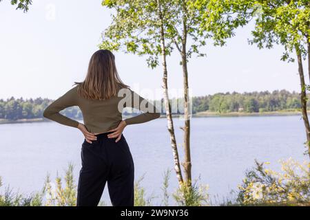 In dieser ruhigen Szene steht eine Frau mit dem Rücken zur Kamera kontemplativ am Rand eines ruhigen Sees. Sie wird von dem zarten Laub von schlanken Bäumen eingerahmt, die zum Teil das ruhige Wasser und den üppigen Wald am fernen Ufer offenbaren. Die Frau trägt ein olivgrünes Oberteil, gepaart mit einer schwarzen Hose, und ihre Hände liegen in einer entspannten Haltung auf dem unteren Rücken. Ihr langes Haar fließt über ihre Schultern und fängt das sanfte Licht eines scheinbar friedlichen Tages ein. Dieses Bild weckt ein Gefühl von Selbstbetrachtung und Ruhe, da die Frau im Denken verloren zu sein scheint oder einfach nur die ruhige Schönheit zu genießen scheint Stockfoto