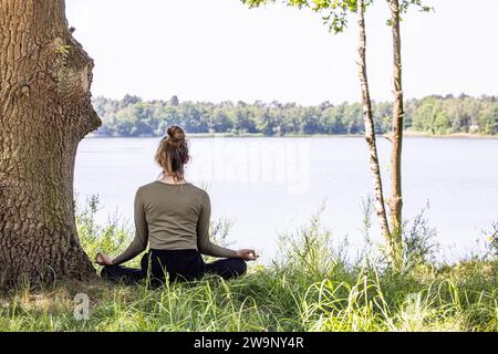 Eine junge Frau findet inneren Frieden, während sie Yoga im Herzen der Natur praktiziert. Sie sitzt anmutig auf dem üppigen grünen Gras neben einem majestätischen Baum und einem ruhigen Waldsee und nimmt an einer Meditationssitzung Teil, die sie mit dem wohltuenden Wesen der natürlichen Welt verbindet. Ruhige Meditation in der Natur. Hochwertige Fotos Stockfoto