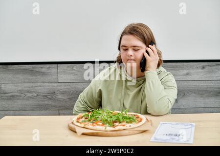 Junge Frau mit psychischen Störungen, die auf dem Smartphone in der Nähe leckerer Pizza in einem modernen gemütlichen Café spricht Stockfoto