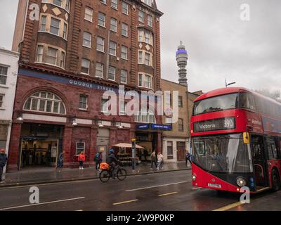 London, Großbritannien. November 2023. Ein klassischer roter Doppeldeckerbus in London fährt an der ikonischen ochsenblutroten Fassade der U-Bahn-Station Goodge Street an der Tottenham Court Road vorbei. Im Hintergrund erhebt sich der BT Tower (früher Post Office Tower) über der Skyline von Fitzrovia. Der vom Architekten Leslie Green entworfene Bahnhof der Northern Line ist ein denkmalgeschütztes Gebäude, das für seinen edwardianischen Barock und seine charakteristischen glasierten Terrakottafliesen berühmt ist. Die Szene fängt das Wesen der Verkehrsinfrastruktur und historischen Architektur im Zentrum Londons an einem nassen Herbstmorgen im West End ein. Stockfoto