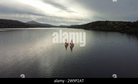 Segeln unter bewölktem Himmel auf Lake Bala, Wales. Gestreifte Segel in leuchtenden Farben sorgen für einen ruhigen Kontrast vor der bergigen Kulisse. Stockfoto